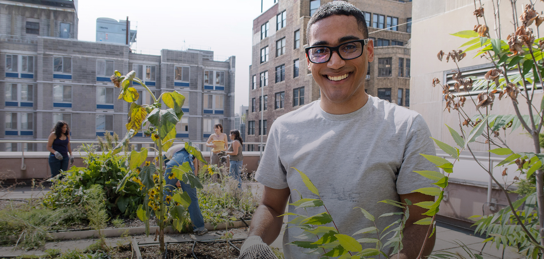 A male student wearing a white t-shirt and black framed glasses is smiling on top of the roof next to the dye garden