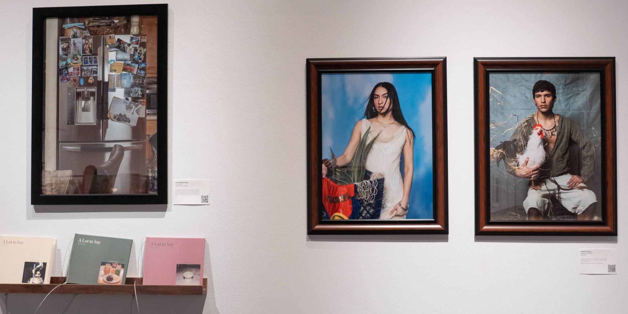 three large photographs on the wall. One photograph is accompanied by an installation of three books on a narrow ledge shelf. In one of the two other photographs appears a seated man holding a rooster, in the second a standing woman with the hand placed on a large vessel with green leaves and bright colored fabric showing.