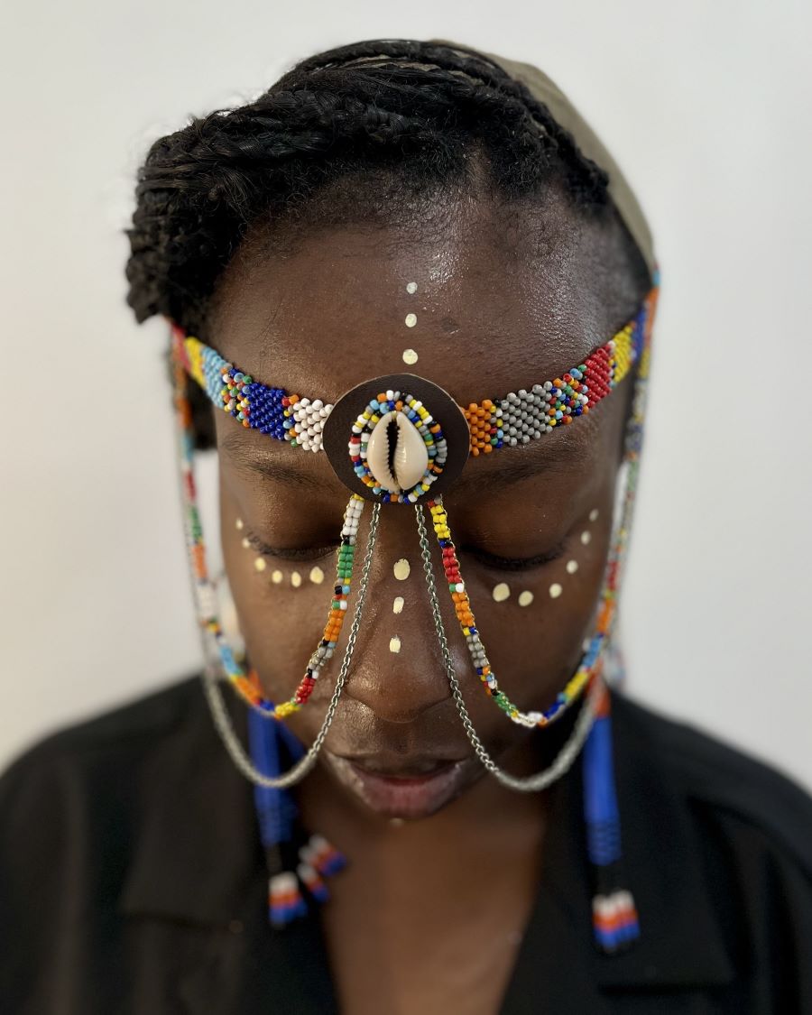 A portrait photograph in color, portraying a female wearing a headdress made of beads, chains, and a shell at the center where the strands meet; dots adorn the eyes, forehead, and nose.