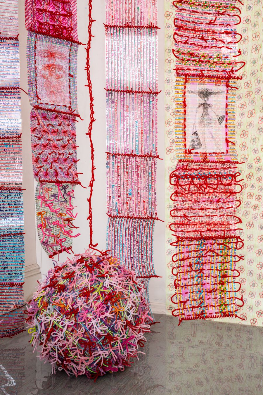 arrangement of space and wall hangings of various widths and lengths made of dura-lar film with interwoven chenille stems. Shades of pink, red, and yellow dominate the installation. Lines of written word in pastel multicolor pens lend additional patterns to the textured surfaces. There are several drawings embedded in several panels. a large ball of chenille stems tied into bows rests on the floor