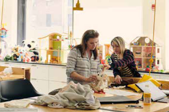 In a brightly lit toy design lab two women are standing at a large work table with fabric, tools, and paper strooned about. One of the women is holding a plush toy, which both are examining. In the background, several doll houses and other toys are displayed on the window sills.