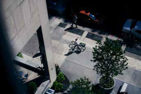 birds eye view looking down the street from somewhere high on the building, the downward lines of the building's wall and columns anchor the composition. It's a warm day, there are few people around, one person is walking the bicycle. There are benches, small trees in large containers, and several cars parked along the street. Sunlit grays of the sidewalk and building concrete and very dark shadows create a slightly unsettling feeling about the scene.