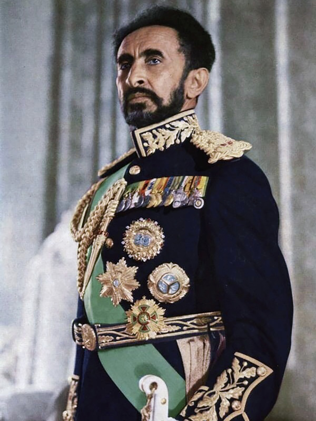 Emperor Selassie I wearing a black military jacket with elaborate gold embroidered details, ribbons, medals, and green sash with ceremonial sword. 