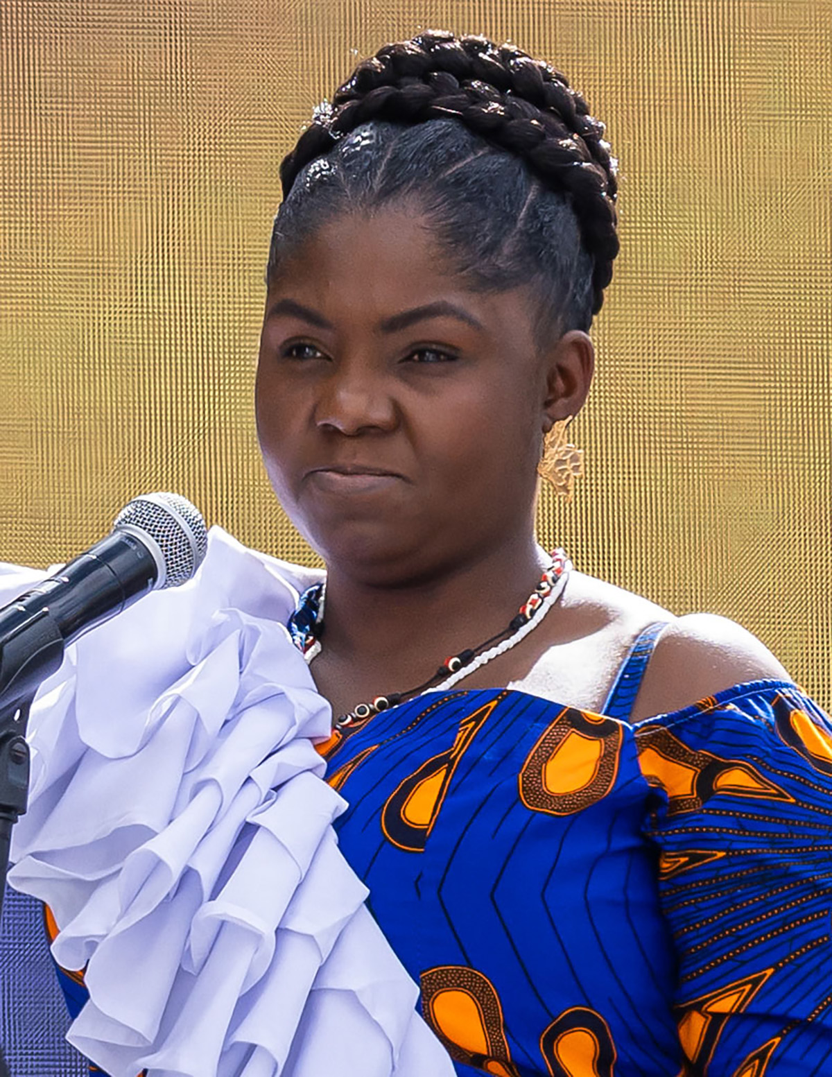 Vice President Francia Márquez (a Black woman), stands at a microphone wearing her hair in braids coiled at the crown of her head with a blue dress. 