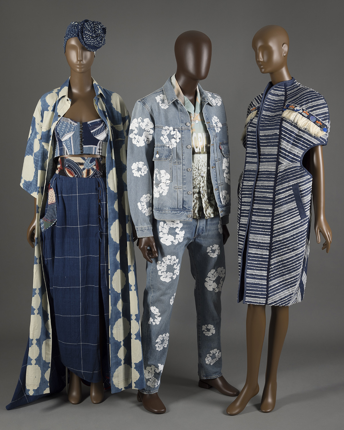  Three brown mannequins dressed in blue ensembles. Left to right: female mannequin dressed in dark blue headwrap, light blue and white floor-length jacket, multi-colored patchwork crop top, and dark blue skirt with white grid pattern. Male mannequin dressed in light blue denim jacket and jeans with a white cotton flower print and light blue printed shirt. Female mannequin dress in a patterned blue denim coat. 