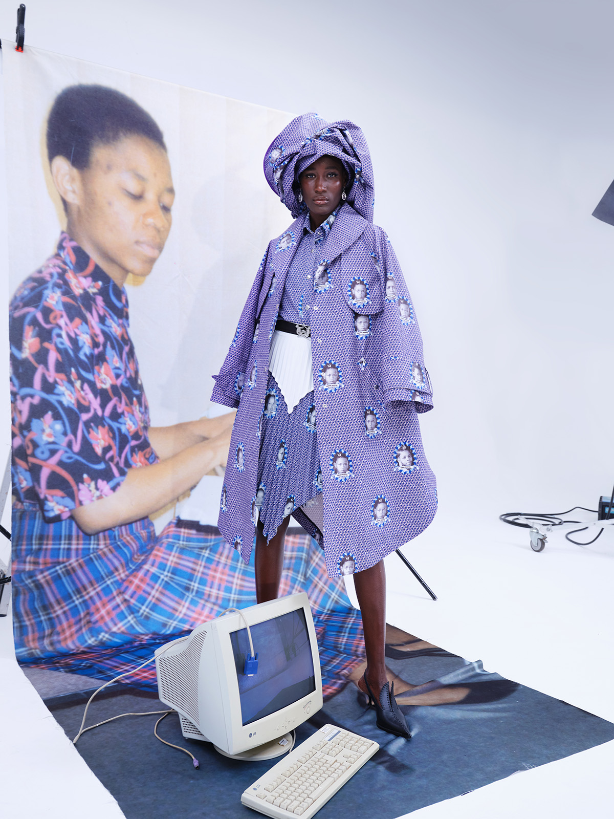 Black woman posing with large photo and computer, wearing purple coat, dress, and hat ensemble printed with images of family members