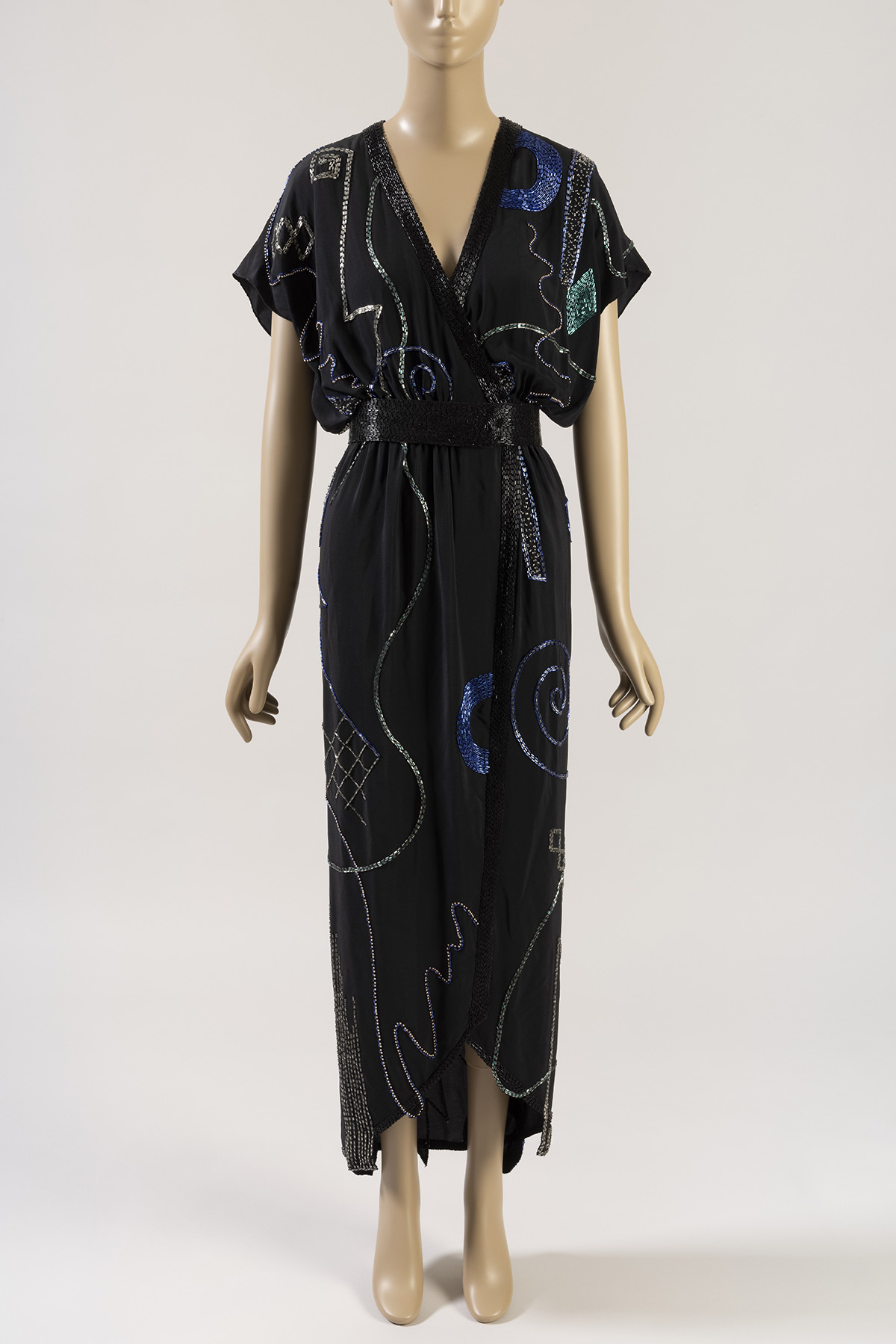 Beige mannequin wearing a V-neck black dress with an uneven hem that reaches to the ankles. It is embroidered with wavy and swirling lines of beads in silver, blue, and green. 