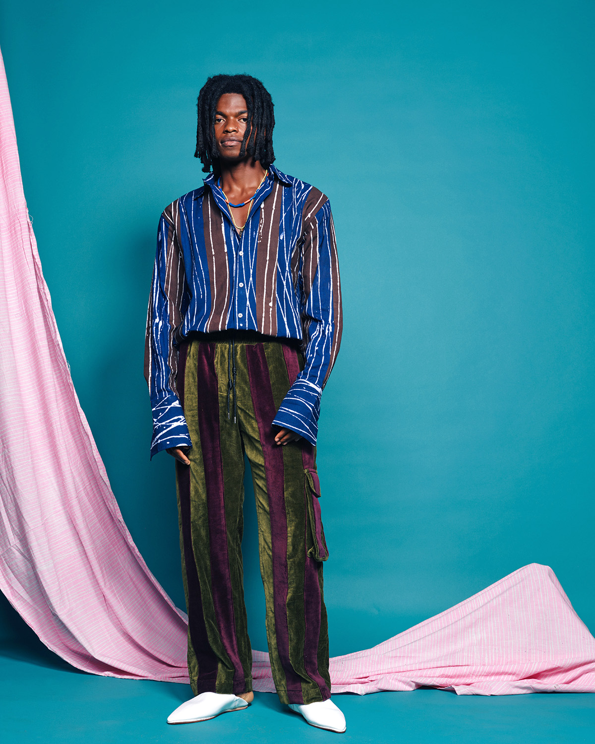 A Black man with chin length dreadlocks wears a brown and blue striped button down shirt, olive green and burgundy striped trousers, and white shoes against a turquoise background with a draped pink cloth.