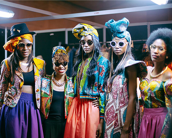 5 stylish Black women wearing pattenered jackets and headscarves with colorful skirts and sunglasses