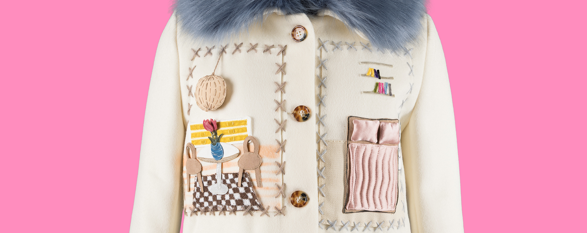 Detail shot of a cream wool coat with a blue faux fur collar, hand-stitched with X-shaped cross-stitching and appliquéd dollhouse-style scenes, including a bookshelf, a dining table with tulip vase and chairs on a checkered rug, and a made bed with pink linens, on a pink background