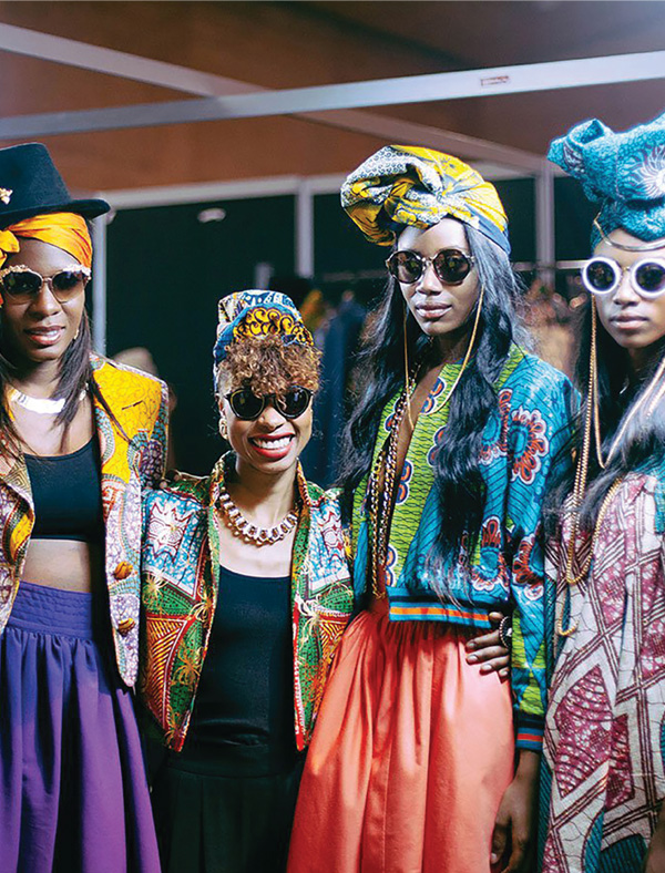 Four stylish Black women wearing pattenered jackets and headscarves with colorful skirts and sunglasses