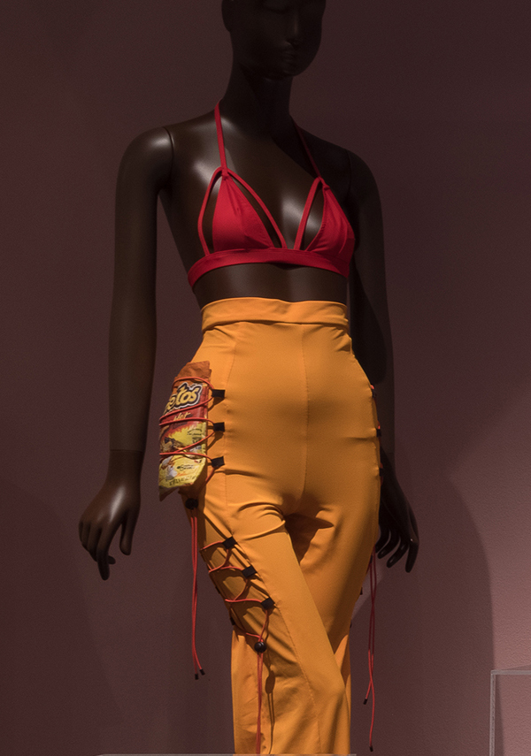 Mannequin wearing halter top with snug pants with lacing on the side of the legs holding a bag of cheetos 